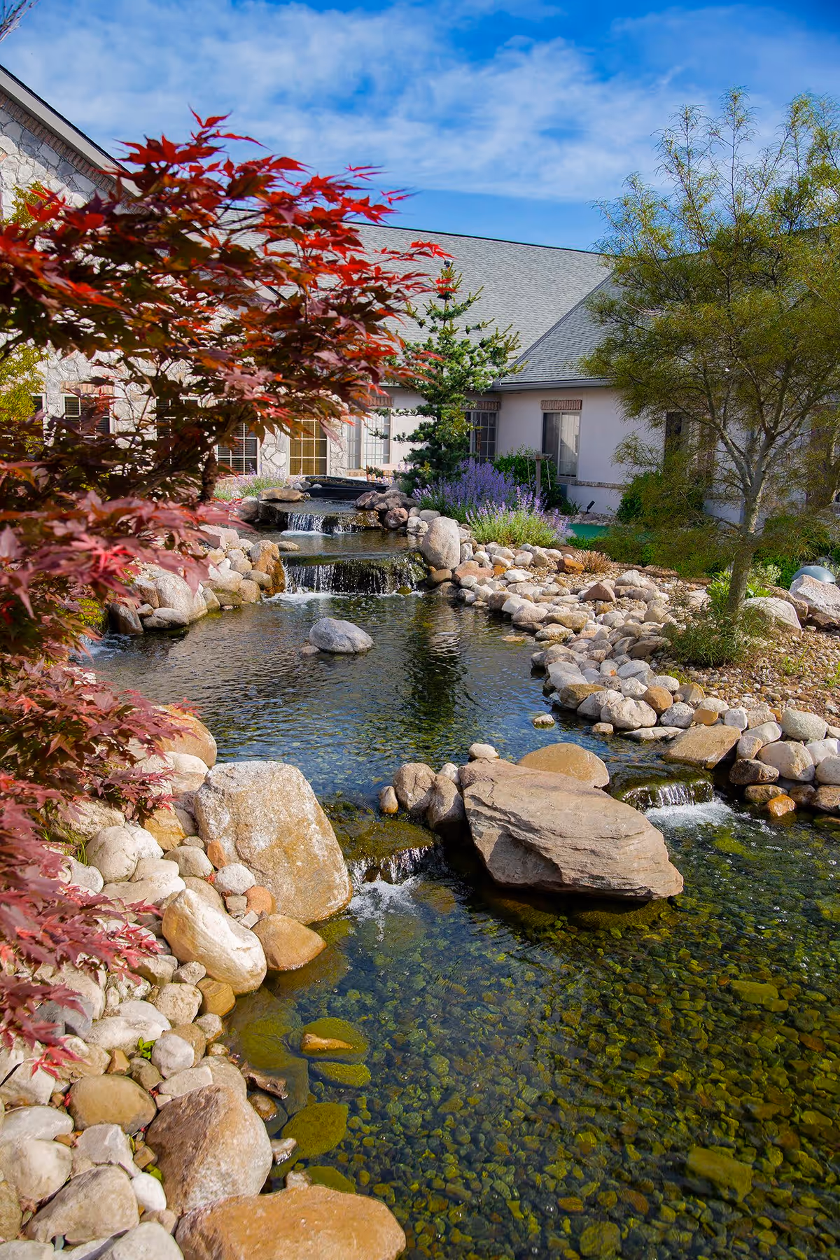 A serene outdoor garden area at The Inn at Winchester Trail featuring a clear water stream with small waterfalls flowing over rocks, surrounded by various plants and trees, with a building visible in the background under a partly cloudy blue sky.