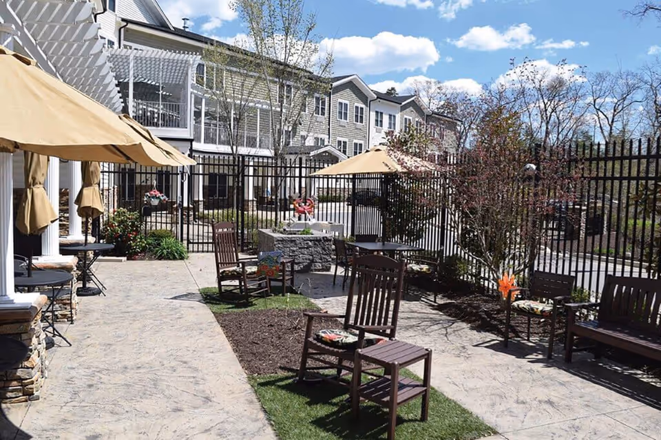 Outdoor patio area at Spring Arbor of Severna Park with several wooden chairs and tables under beige umbrellas, surrounded by a black metal fence and landscaping with trees and bushes. The building with multiple windows is visible in the background under a partly cloudy sky.