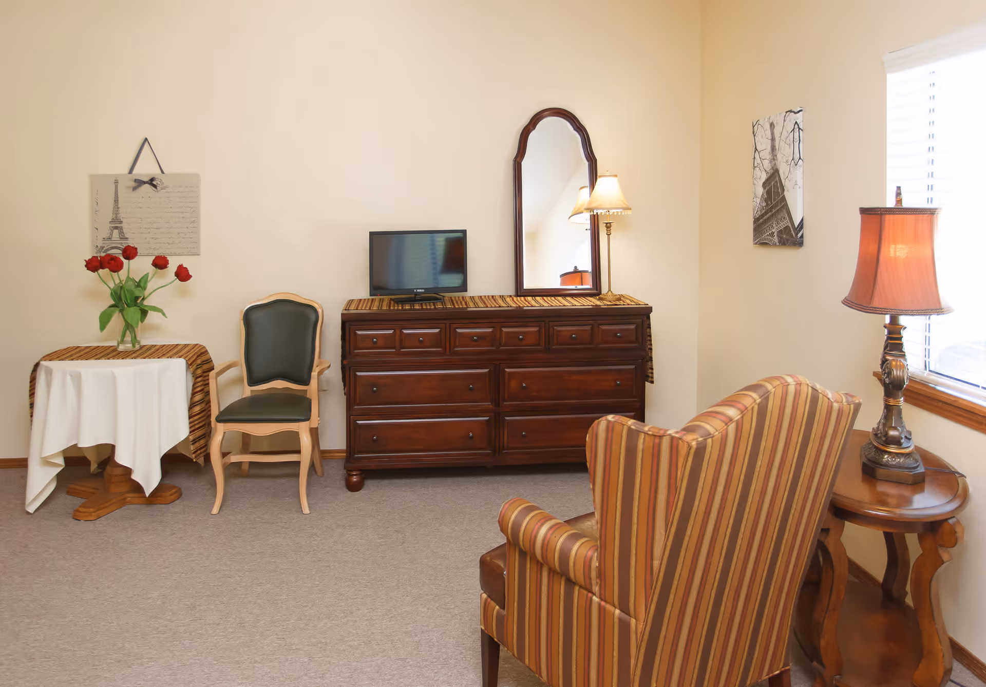 Cozy sitting room with a striped armchair, wooden dresser with mirror and small table holding a vase of red flowers.