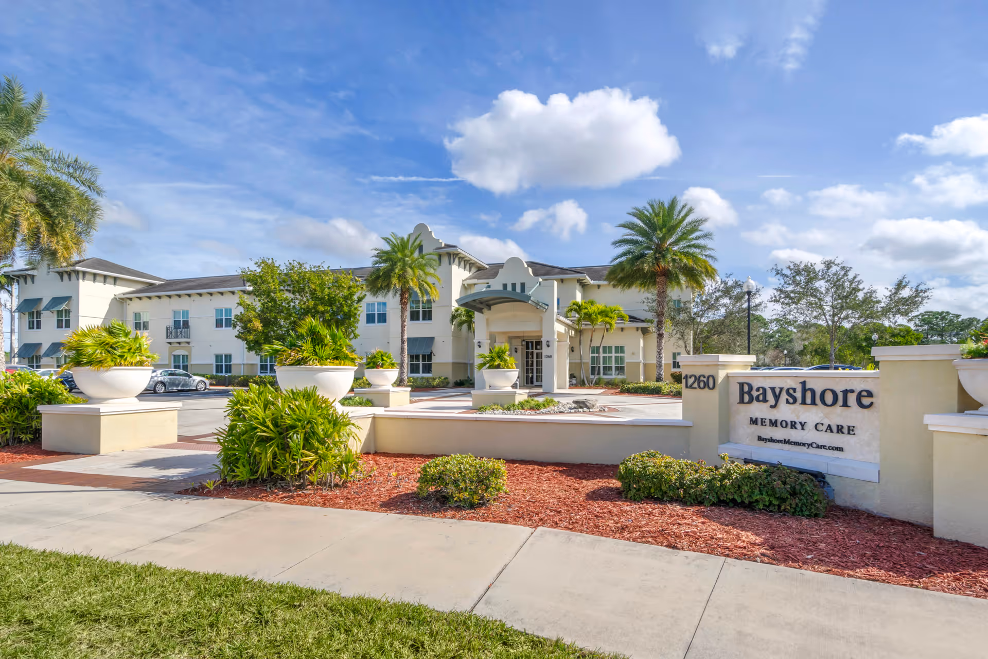 Exterior view of Bayshore Memory Care facility with a clear blue sky, palm trees, and landscaped greenery. The building is two stories with multiple windows and a covered entrance. A sign in front displays the name 'Bayshore Memory Care' and the address number 1260.