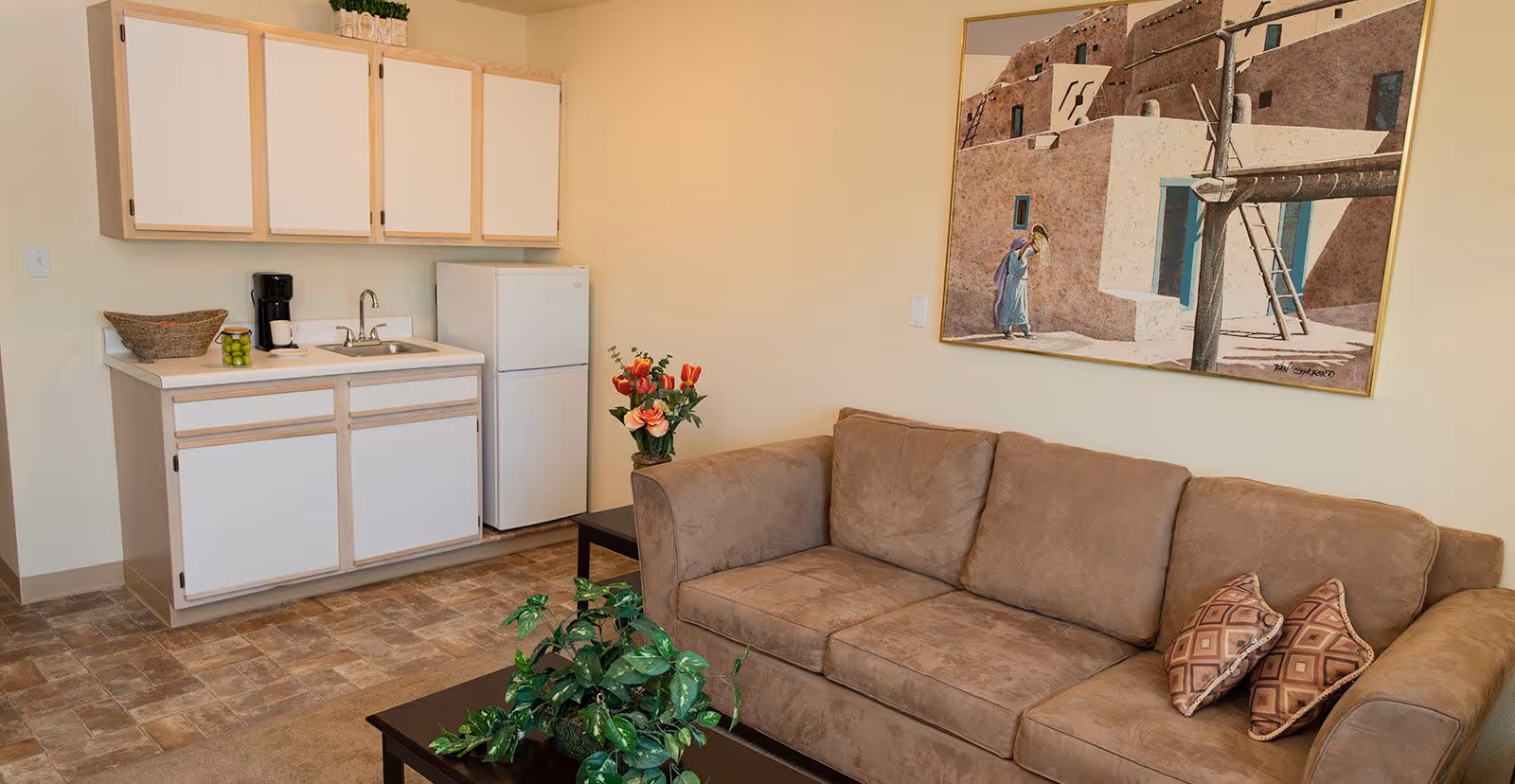 A small living area with a brown couch adorned with two patterned pillows, a coffee table with a green plant, and a kitchenette featuring white cabinets, a sink, a coffee maker, and a refrigerator. A painting depicting a southwestern adobe scene hangs on the wall above the couch.