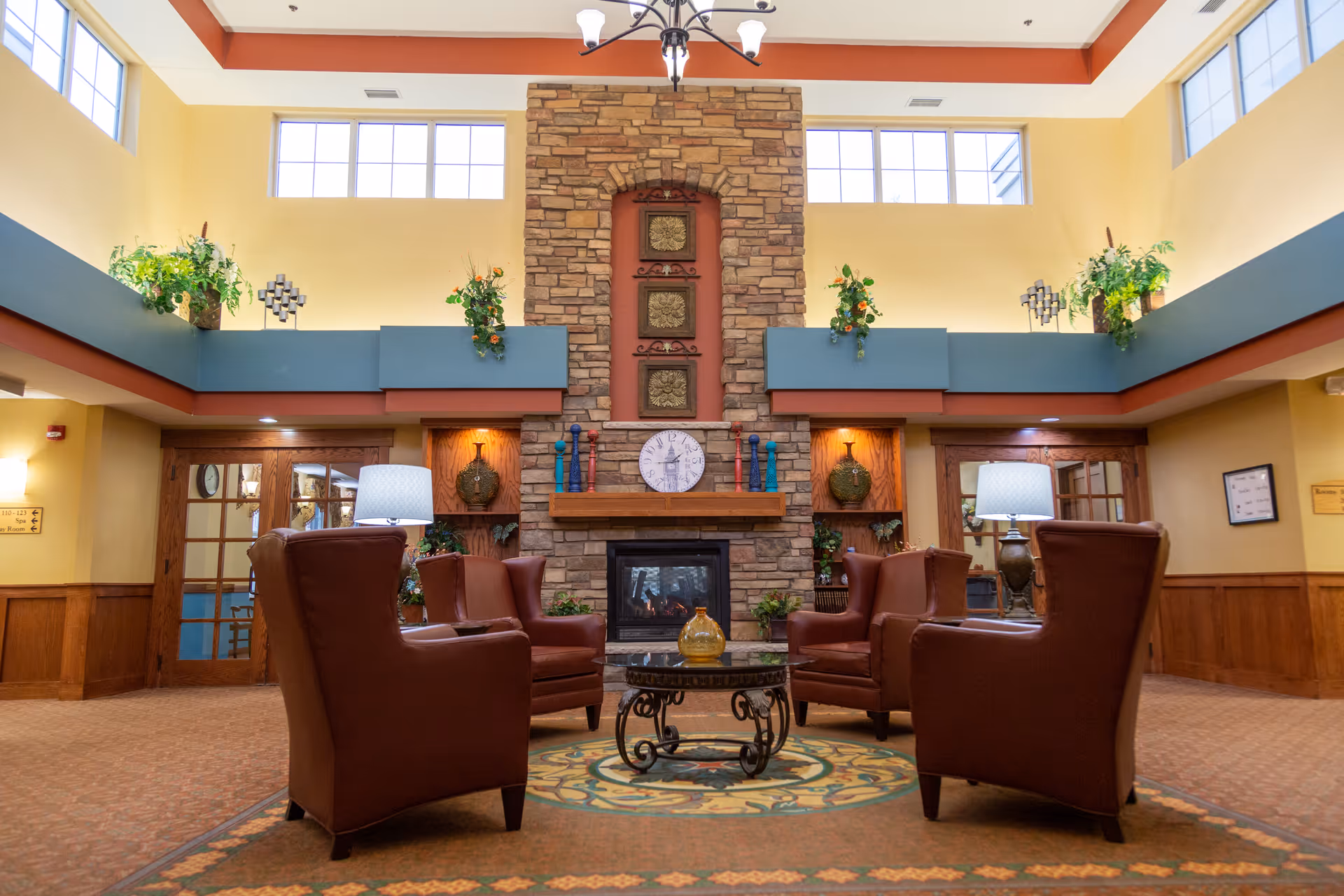 A cozy senior living facility common area with four brown leather armchairs arranged around a round glass coffee table on a patterned rug. The room features a tall stone fireplace with decorative items and a clock above the mantel. There are large windows near the ceiling allowing natural light, plants on ledges, and warm yellow walls with wood paneling on the lower half.