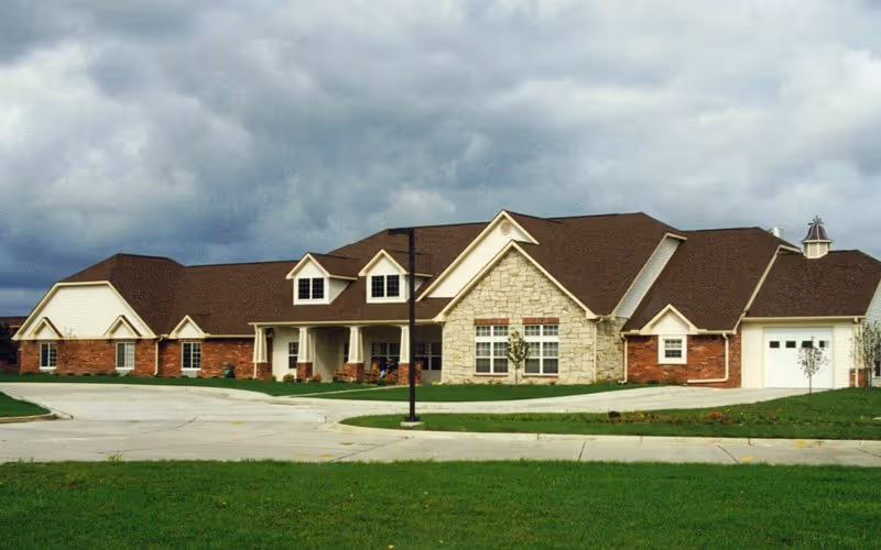 Front exterior of a single-story brick-and-stone senior living building with a circular driveway, lawn, and cloudy sky.