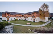 Exterior view of a two-story senior living facility building with white walls and wooden accents. The building surrounds a landscaped courtyard with grass, benches, a tree, and a paved walkway. The sky is clear and the setting appears calm and inviting.
