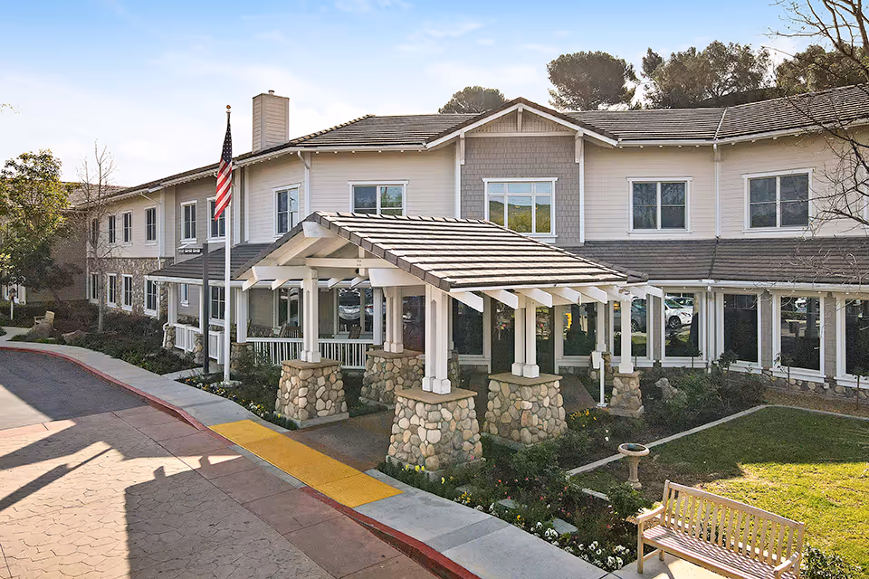 Front entrance of a two-story residential facility featuring a covered stone-column porte-cochère, flagpole, bench, and landscaped lawn.
