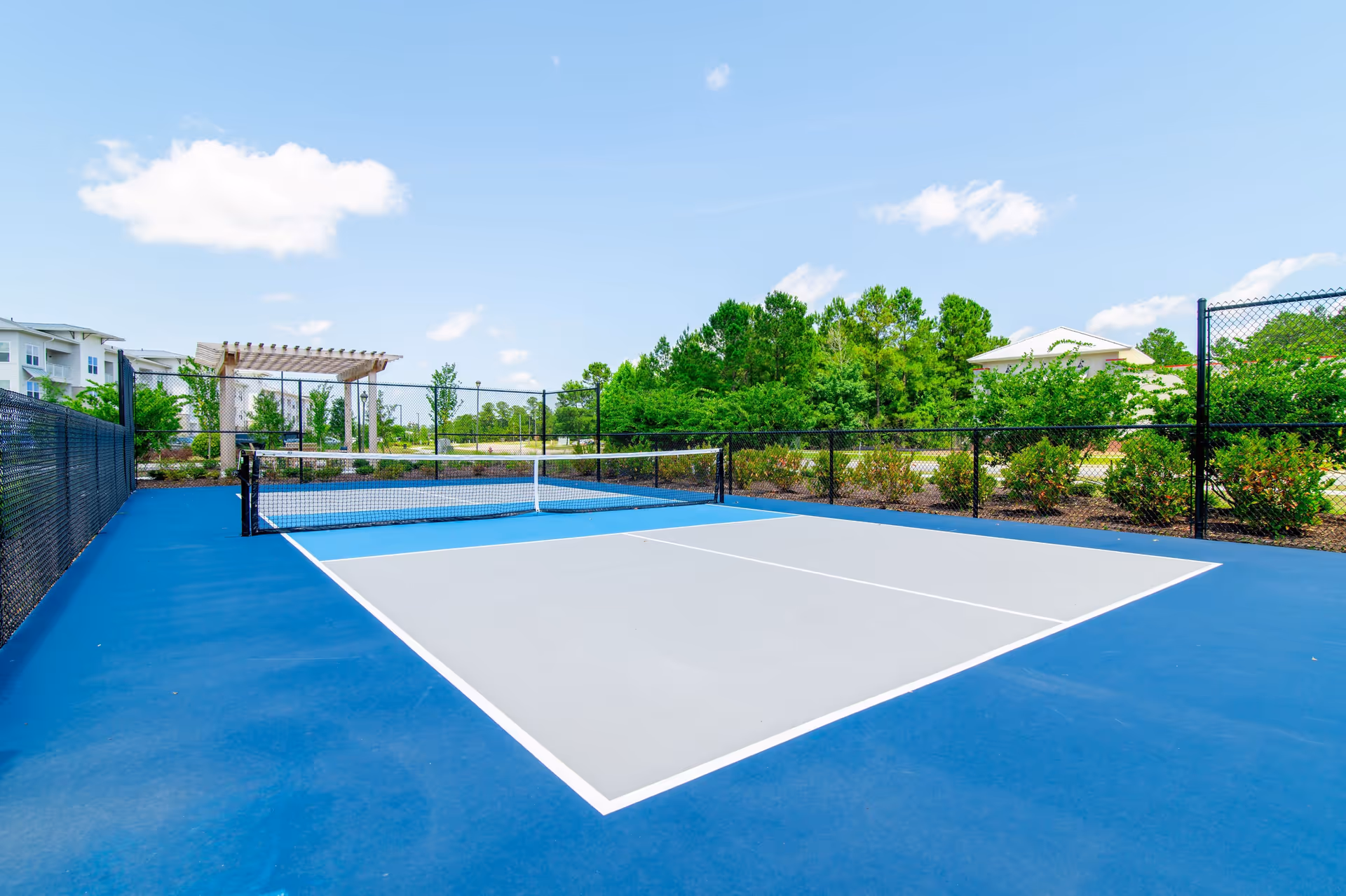 Outdoor pickleball court with blue and gray surface, surrounded by a black chain-link fence. In the background, there are green trees, shrubs, a pergola, and residential buildings under a blue sky with some clouds.
