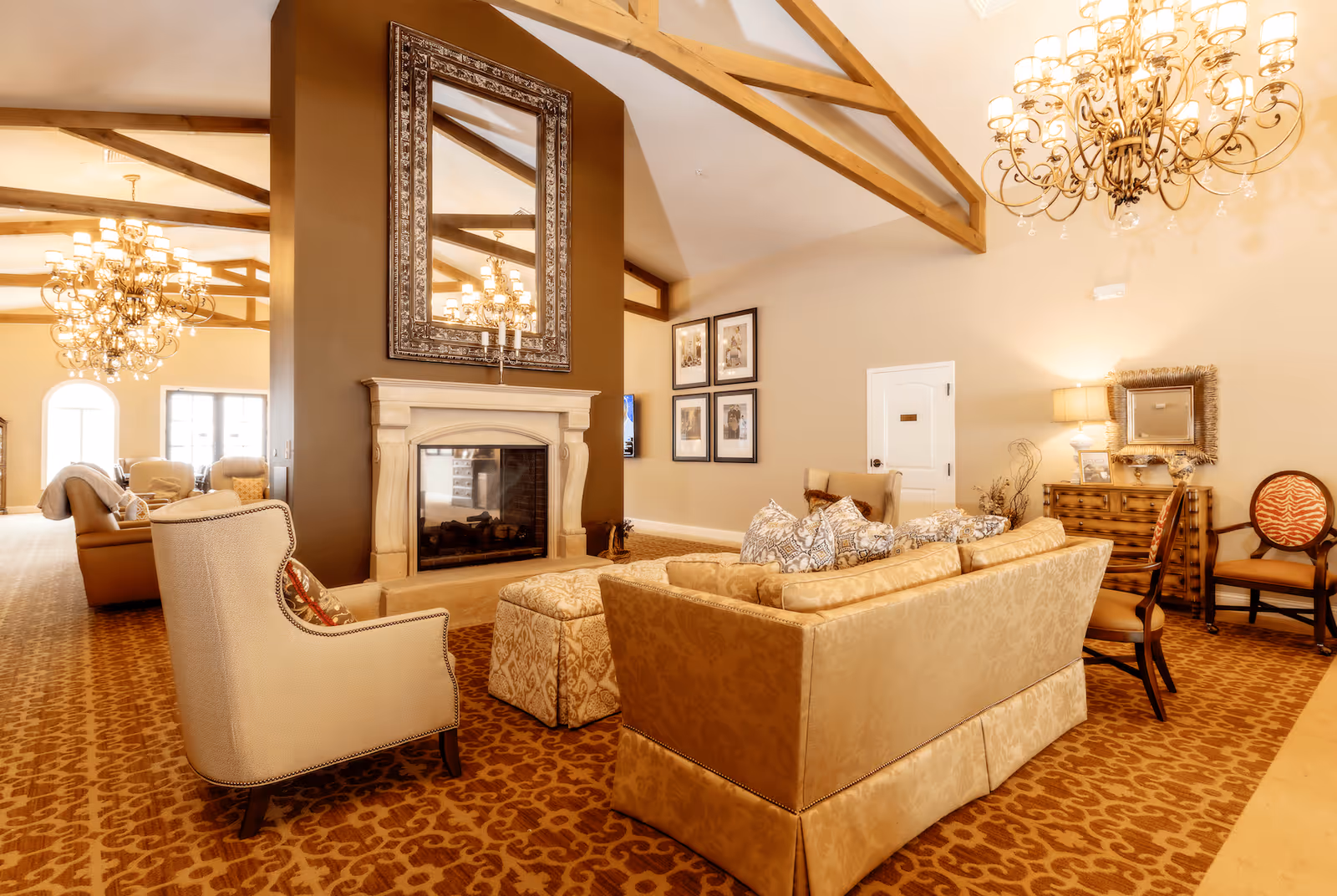 A warm and elegant living room area with a large beige sofa adorned with patterned pillows, a beige armchair, and two ottomans. The room features a stone fireplace with a large ornate mirror above it, exposed wooden ceiling beams, and two large chandeliers. The walls are decorated with framed pictures and a mirror above a wooden dresser. The carpet has a patterned design in shades of brown and beige.