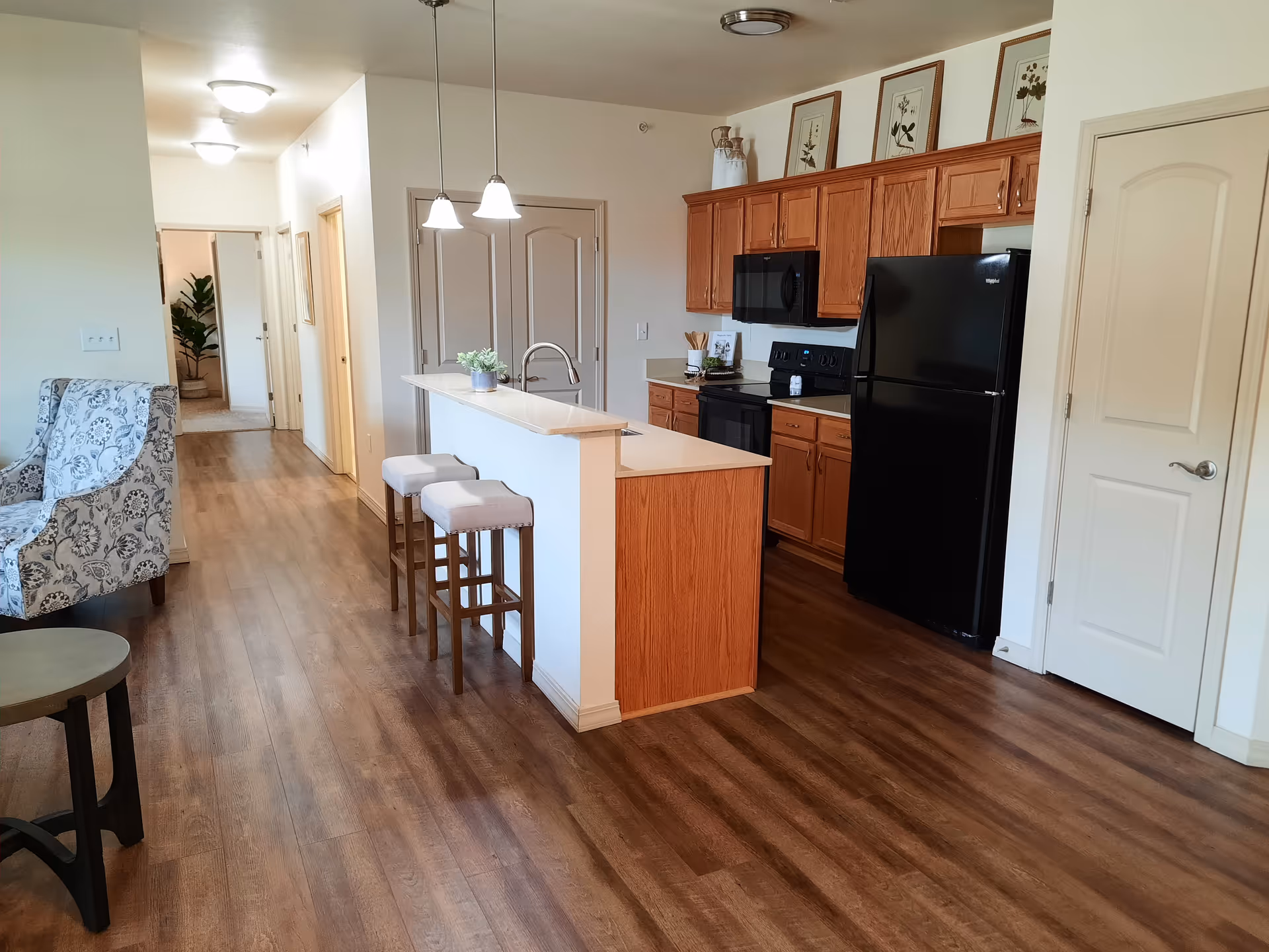 Interior view of a senior living facility kitchen and hallway. The kitchen features wooden cabinets, a black refrigerator, stove, and microwave, a white countertop island with two cushioned bar stools, and pendant lights hanging above the island. The hallway leads to other rooms, with a patterned armchair and small table visible on the left side.