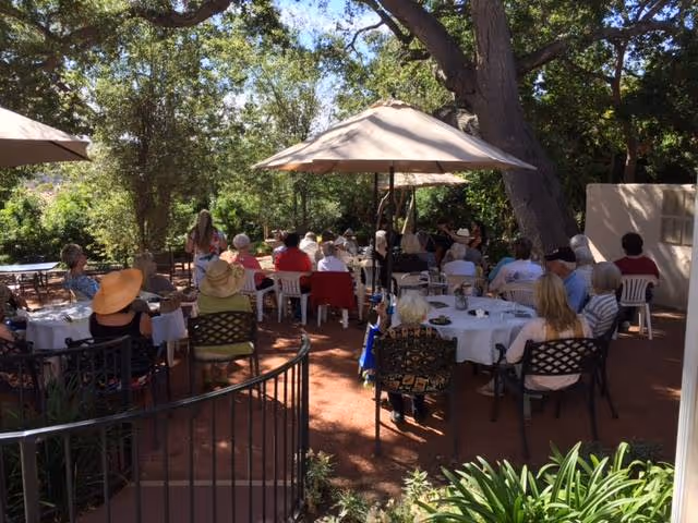 Seniors seated at round tables under umbrellas on a tree-shaded outdoor patio during a gathering.