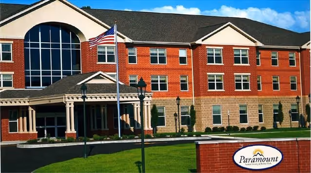 Front exterior of a three-story red-brick senior living building with an American flag and a Paramount sign on the lawn.