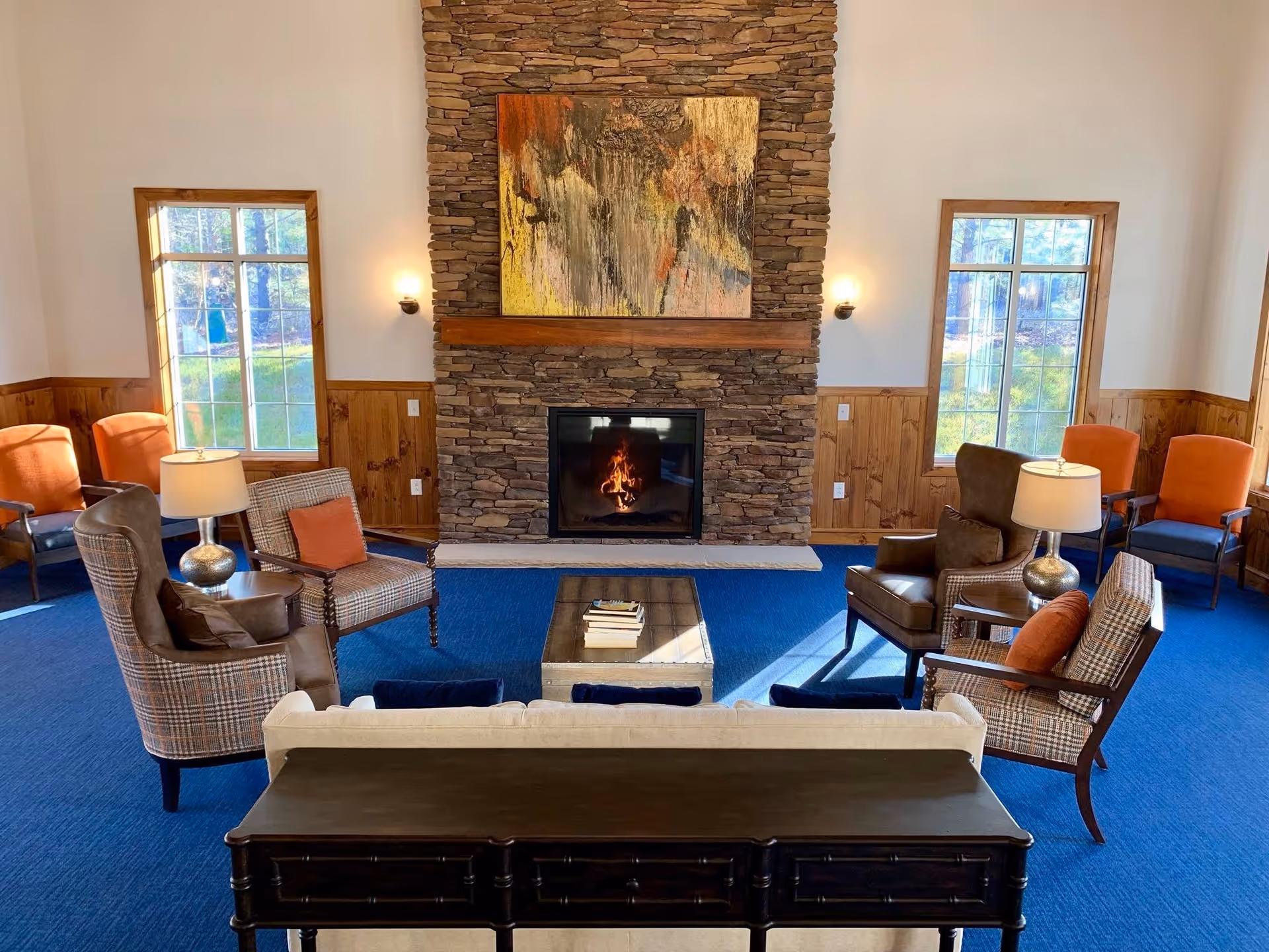 A cozy living room with a stone fireplace in the center, a colorful abstract painting above it, and two windows on either side letting in natural light. The room features a blue carpet, wooden paneling on the lower walls, and a variety of seating including armchairs with cushions, a sofa, and side tables with lamps.