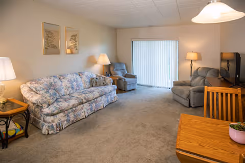 A living room with a floral patterned sofa, two gray recliner chairs, a wooden dining table with chairs, a TV on a stand, and three lamps providing warm lighting. There are two framed pictures on the wall and vertical blinds covering a sliding glass door.
