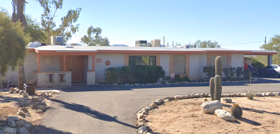 Single-story building with a flat roof and a covered entrance, surrounded by desert landscaping including cacti and rocks, under a clear blue sky.
