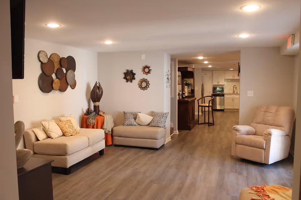 A cozy living room area with beige cushioned chairs and sofas, decorative pillows, wall art including circular woven pieces and sunburst mirrors, a small table with a decorative sculpture and blankets, and a view into a kitchen area with white cabinets and stainless steel appliances.