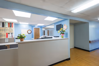 Interior view of a healthcare facility reception area with a white counter, potted plants, a wall clock, file organizers, and a hallway leading to other rooms. The walls are painted light blue and the floor has wood-like flooring.