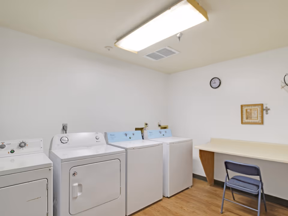 Small communal laundry room with multiple washers and dryers, a folding table and a chair under fluorescent lighting.