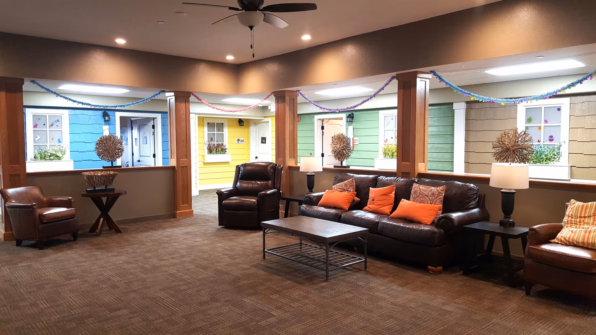 A cozy common area with brown leather chairs and a sofa adorned with orange and patterned pillows. The room has a ceiling fan and recessed lighting. Behind the seating area, there are colorful cottage-style doors and windows painted blue, yellow, green, and tan, decorated with flowers and festive garlands hanging across the ceiling.