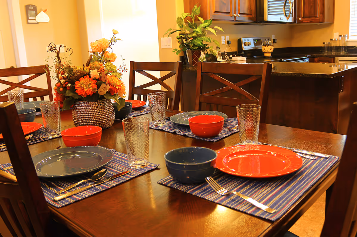 A dining table set for four with blue and red plates and bowls, clear textured drinking glasses, silverware, and blue striped placemats. A floral centerpiece with orange and yellow flowers is in the middle of the table. In the background, a kitchen area with wooden cabinets, a stove, and a countertop is visible.