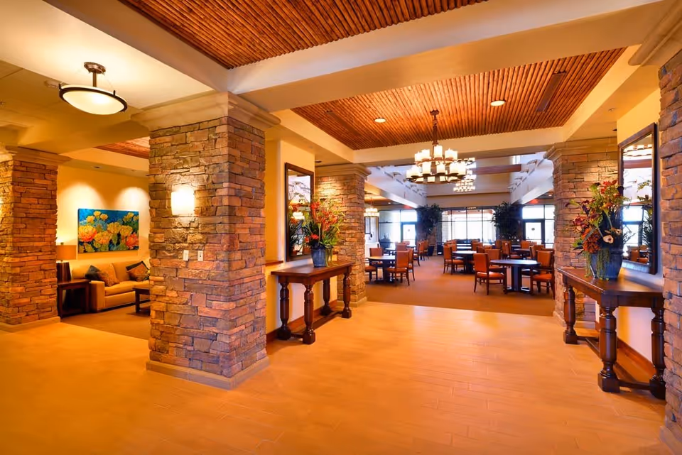 A spacious and warmly lit interior of a senior living facility featuring stone pillars, wooden ceiling accents, and a dining area with multiple tables and chairs. There is a cozy seating area with a couch and colorful floral artwork on the wall to the left, and decorative flower arrangements on wooden tables along the hallway.