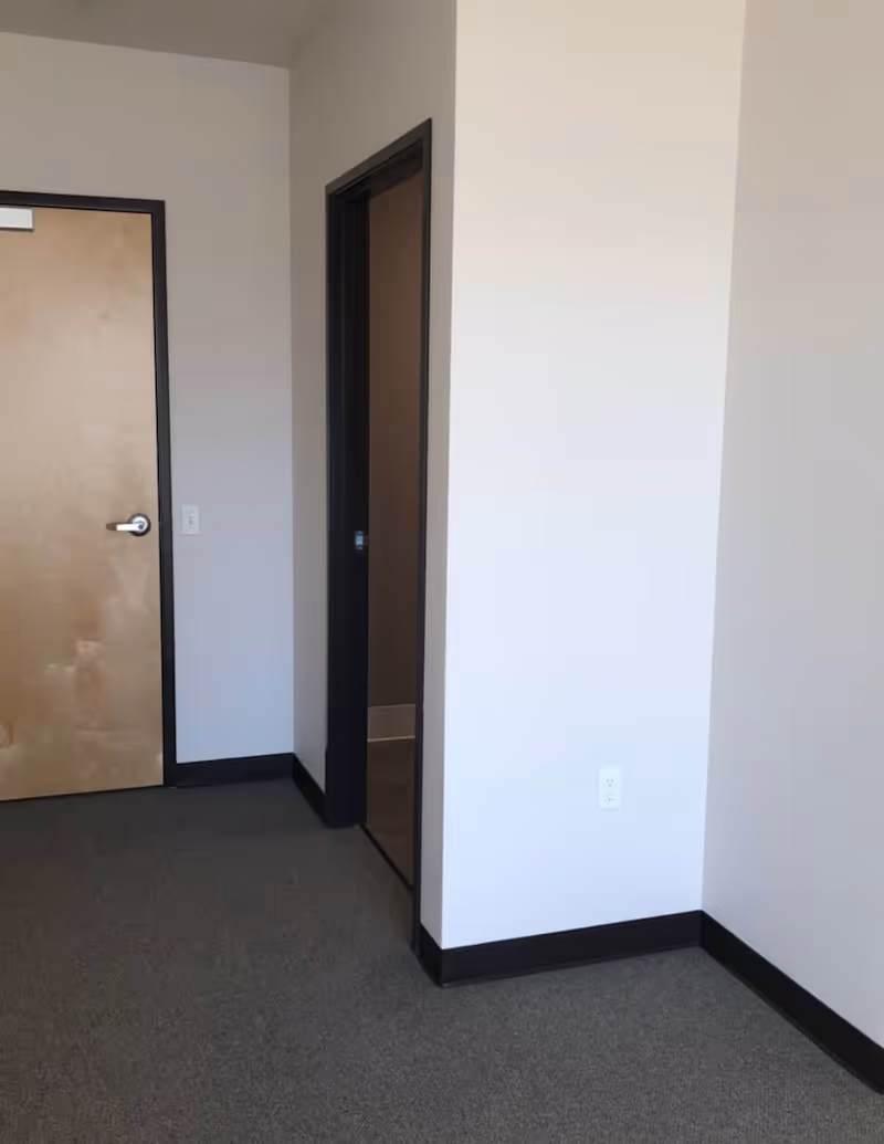 Empty carpeted interior corner showing a closed wooden door, an open doorway, white walls and black baseboards.