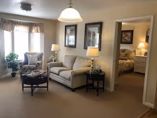 A cozy living room area with a beige loveseat, a cushioned armchair, a round coffee table with magazines, two side tables with lamps, and framed artwork on the walls. There is a doorway leading to a bedroom with a bed and nightstand visible. A window with curtains allows natural light into the room.