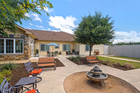 Outdoor courtyard area at Poet's Walk San Antonio, featuring a circular water fountain in the center, surrounded by benches and chairs with red cushions. The courtyard is paved with concrete walkways and landscaped with plants and a tree. The building exterior is visible with beige siding, stone accents, windows, and a patio umbrella over a table with chairs.