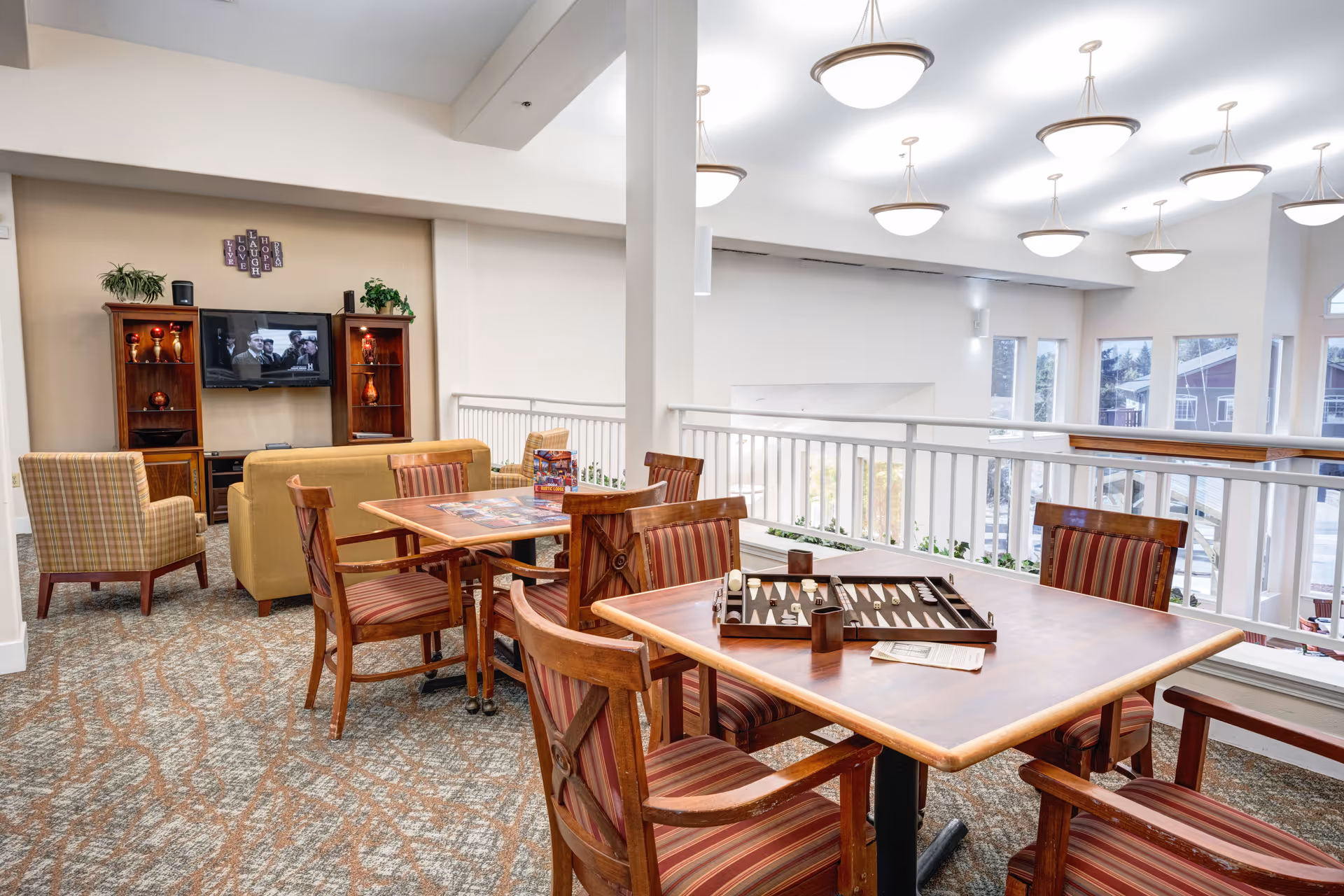 A bright and spacious common area in a retirement facility featuring a seating area with a TV mounted on the wall, two armchairs, and a sofa. In the foreground, there are two wooden tables with striped cushioned chairs around them, one table has a backgammon game set up. Large windows allow natural light to fill the room, and modern ceiling lights hang from the high ceiling.