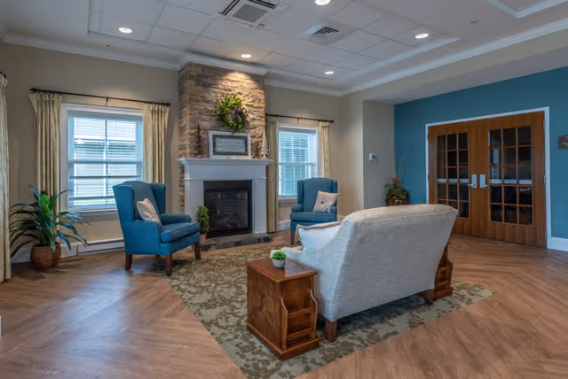 A cozy assisted-living common room featuring a fireplace flanked by windows, two blue armchairs, a sofa on a patterned rug, and wooden double doors.