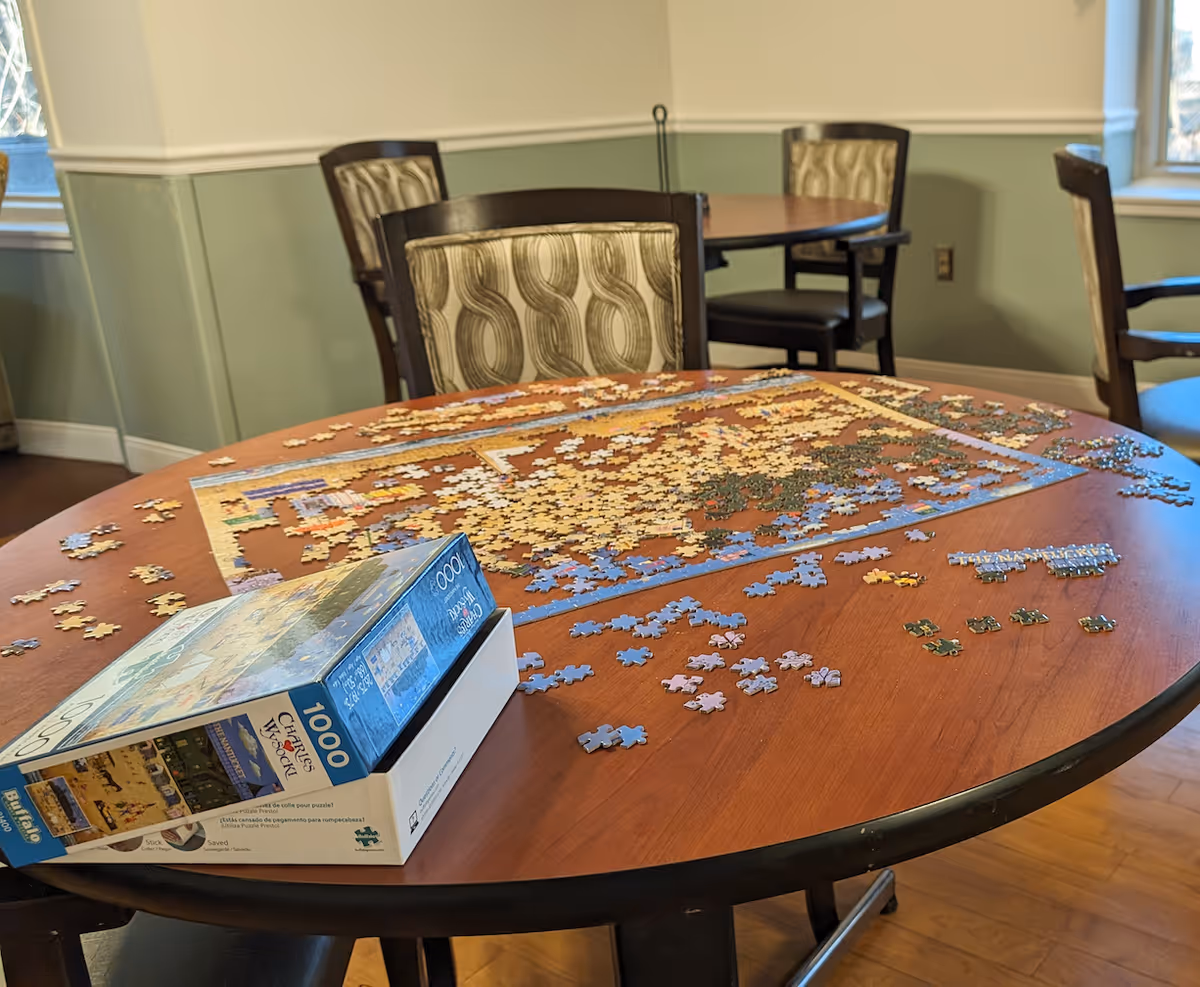 A round wooden table with a partially completed 1000-piece jigsaw puzzle and scattered puzzle pieces. The table is in a room with green and white walls, wooden flooring, and several chairs with patterned upholstery. Sunlight is coming through windows in the background.