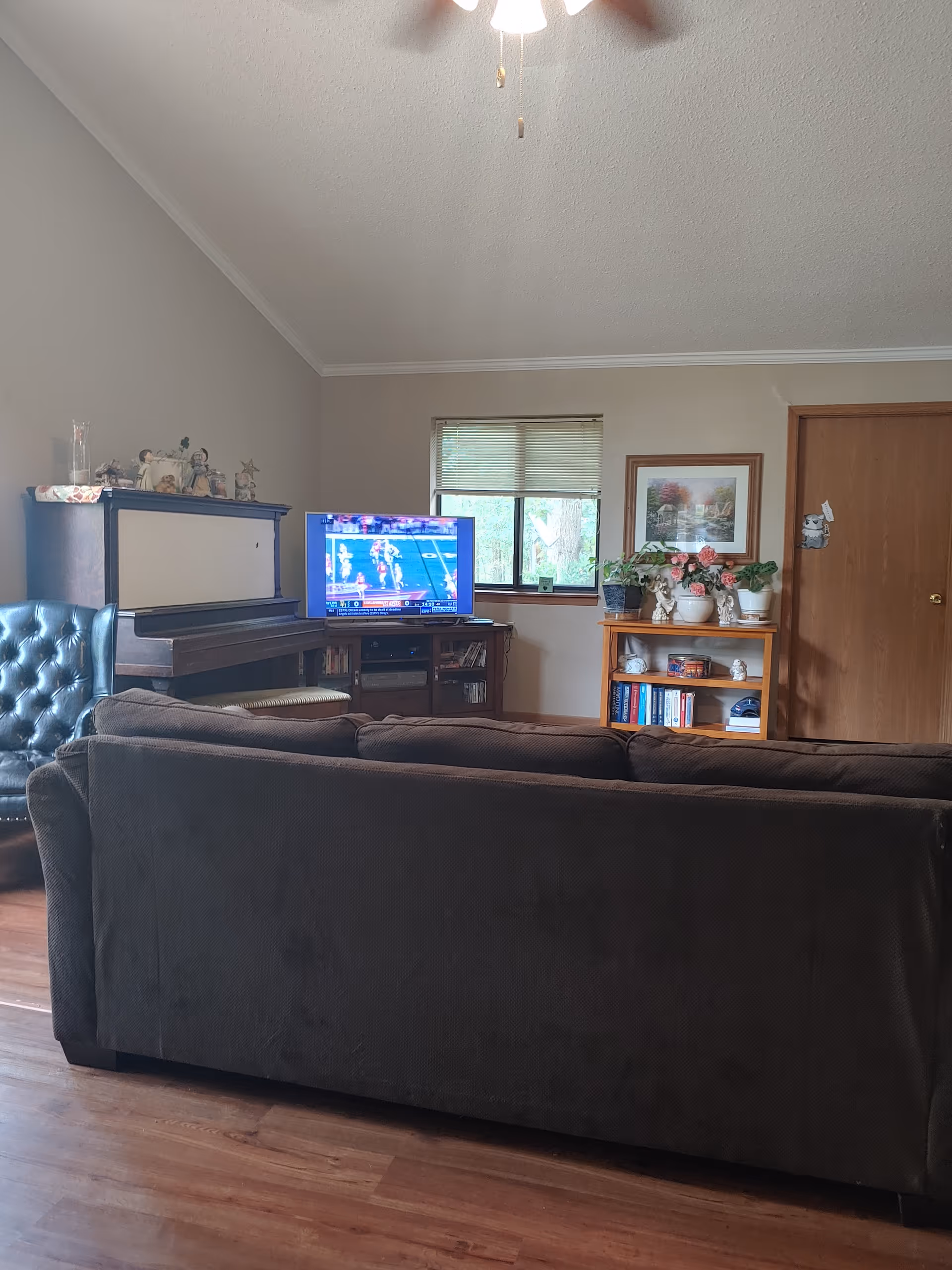 A cozy living room with a brown sofa facing a TV that is showing a sports game. To the left of the TV is a piano with decorative items on top, and to the right is a wooden bookshelf with plants, books, and decorative pieces. A window with blinds is centered on the back wall, and a ceiling fan is partially visible at the top.