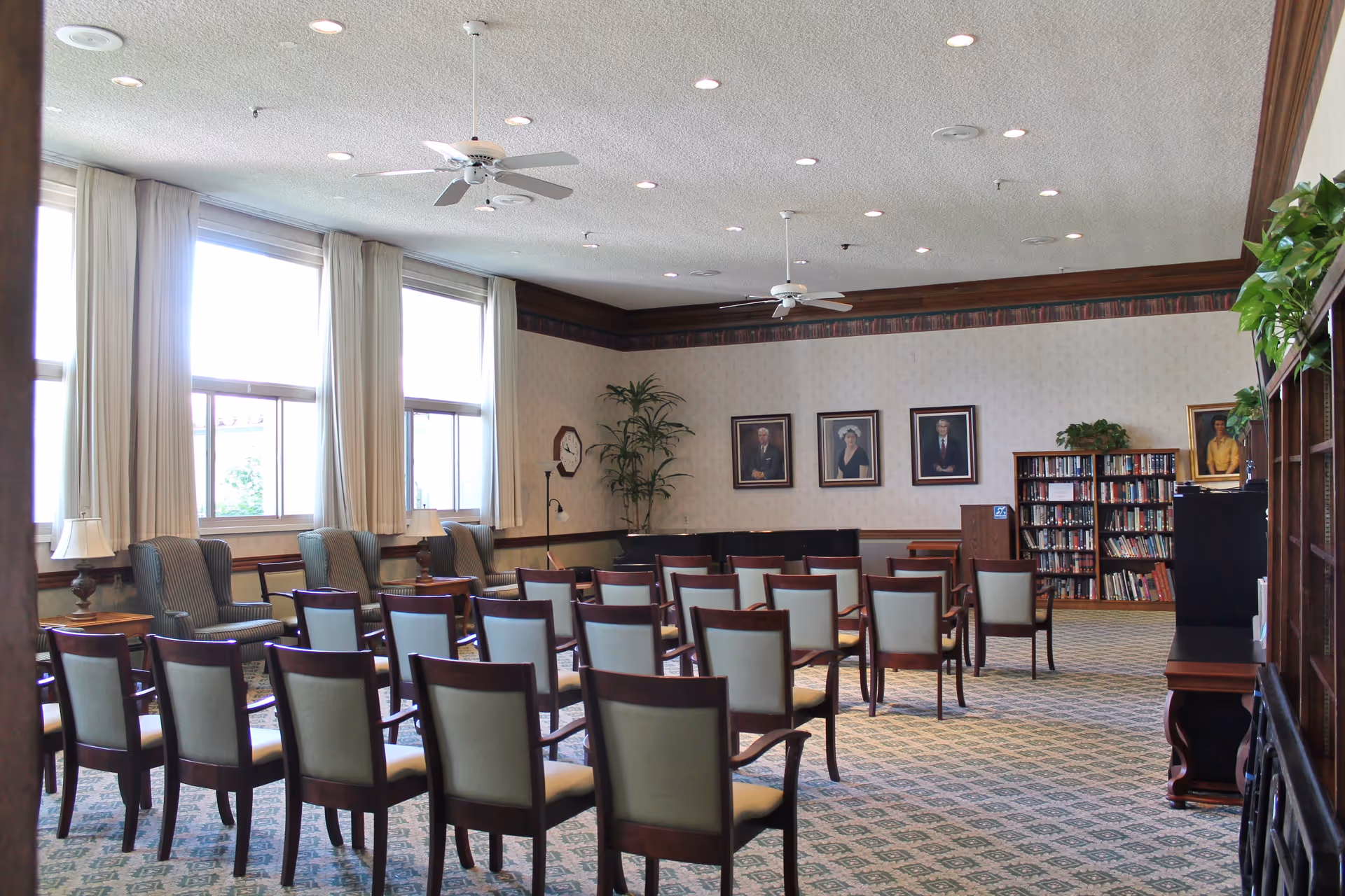A well-lit room with rows of wooden chairs with cushioned seats arranged facing a corner with a piano, bookshelves, and framed portraits on the wall. The room has large windows with curtains, ceiling fans, and a patterned carpet.