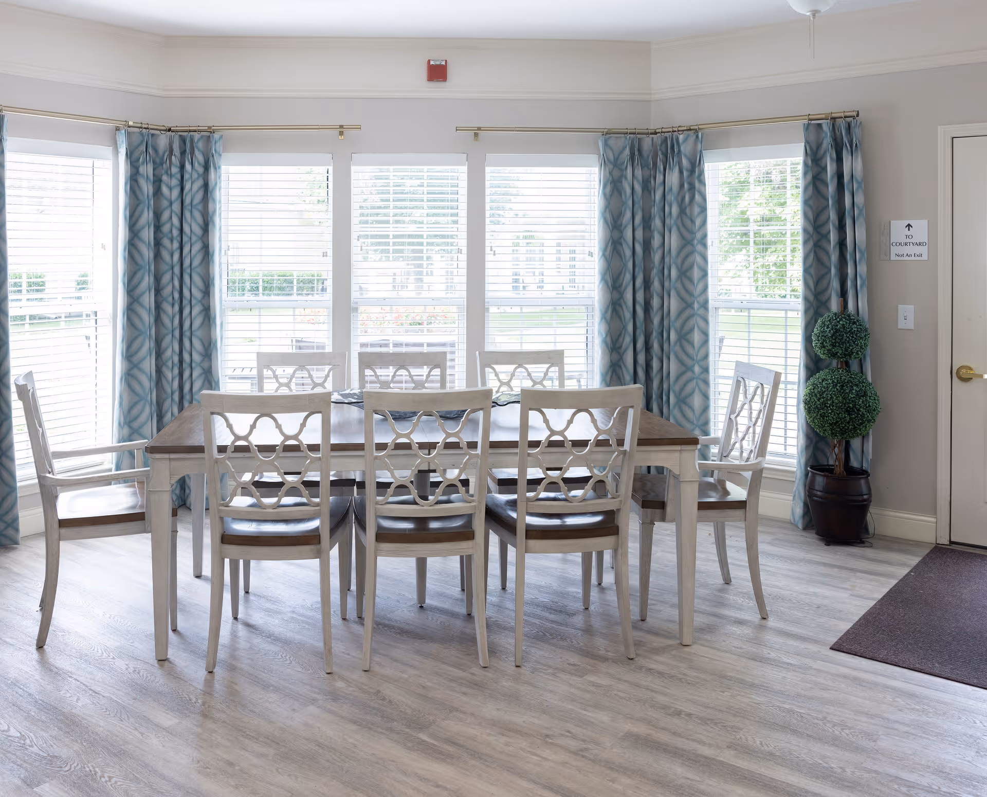 A bright dining room with a wooden table surrounded by eight chairs. The room has large windows with white blinds and blue patterned curtains. There is a potted plant with two green topiary balls near a door with a sign that reads 'To Courtyard Not An Exit'. The floor is light-colored wood.
