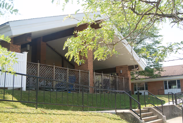 Exterior view of a brick nursing facility's covered entrance with columns, a metal railing ramp and stairs and trees in the foreground.