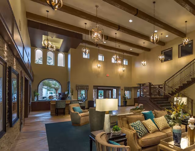 Spacious and well-lit senior living facility common area with high ceilings, exposed wooden beams, multiple hanging light fixtures, comfortable seating including sofas and armchairs with cushions, a large rug, decorative plants, and a staircase leading to an upper floor. Large windows and doors allow natural light to enter the room.