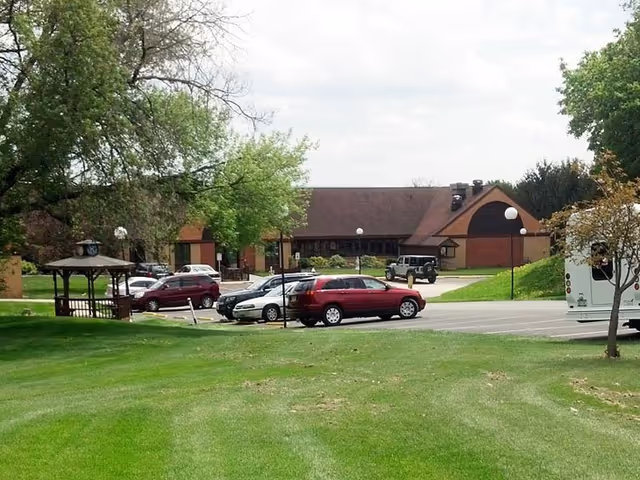 A parking lot with several cars parked in front of a single-story building with a brown roof and brick walls. There is a grassy area with trees and a small wooden gazebo on the left side of the image. The sky is partly cloudy.
