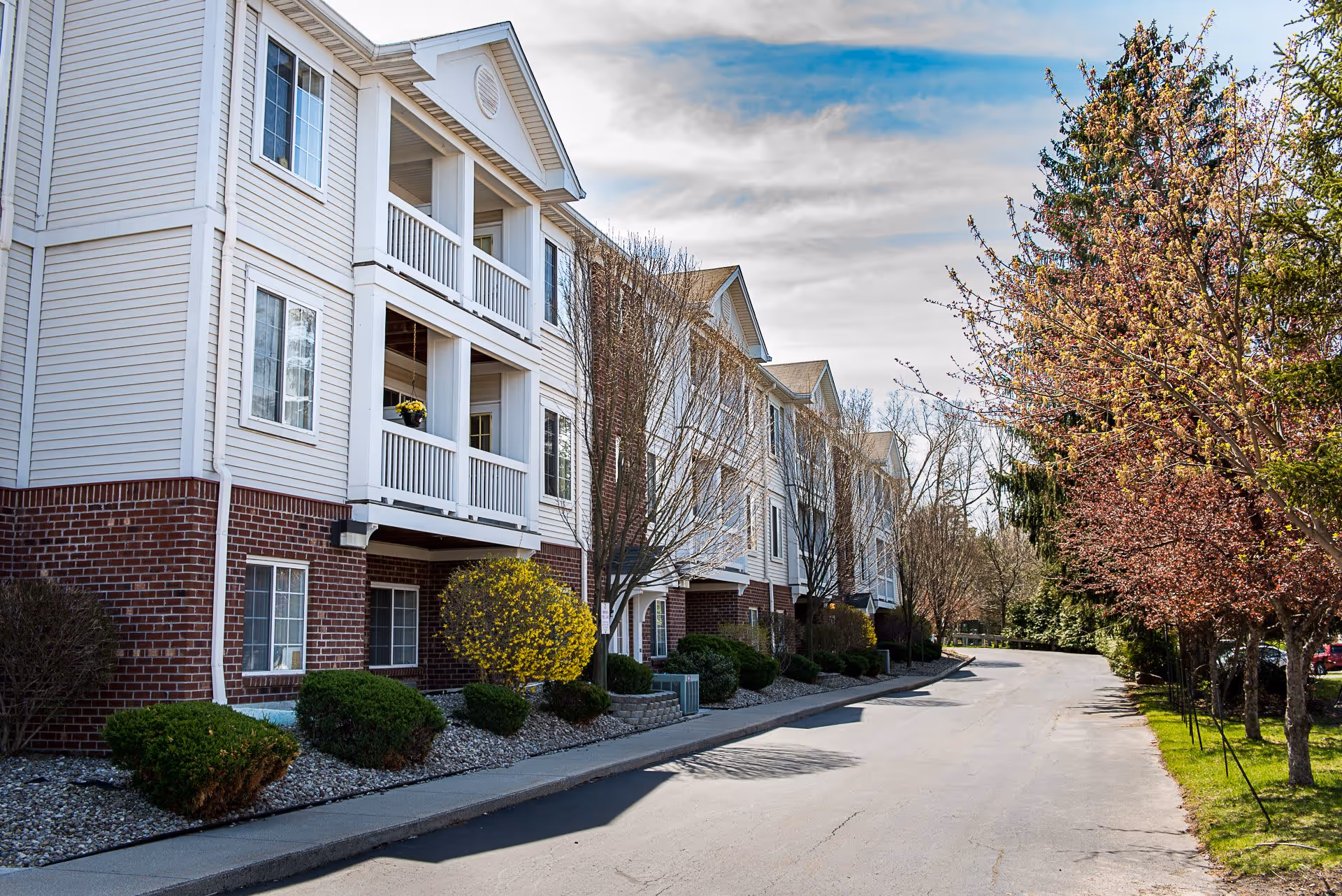 Exterior view of a multi-story senior living facility building with white siding and red brick accents. The building has balconies with railings and is surrounded by landscaped bushes and trees, some with spring blossoms. A paved driveway runs alongside the building under a partly cloudy sky.