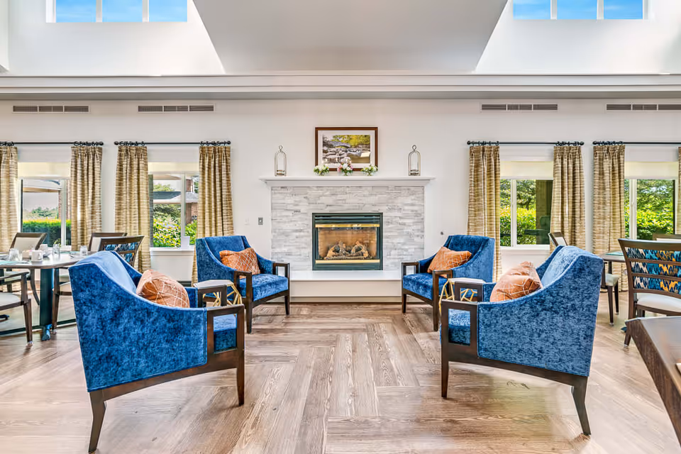 A bright and spacious senior living common area featuring four blue upholstered armchairs with orange accent pillows arranged around a modern gas fireplace with a white stone surround. Large windows with beige curtains let in natural light, and there are dining tables and chairs visible in the background. The room has a wooden floor and a high ceiling with skylights.