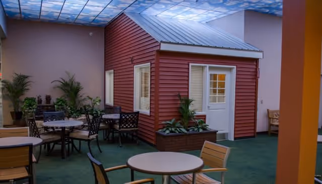 Indoor common area with round tables and chairs surrounding a small red house-like structure, potted plants, and a skylight-style ceiling.