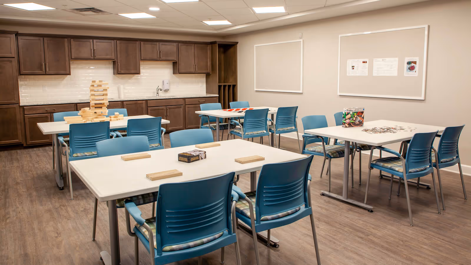 A well-lit activity room with several white tables and blue chairs arranged around them. On the tables are various games including a large wooden block stacking game, a puzzle, and a checkers set. The room has wooden cabinets and a sink along one wall, with two bulletin boards on another wall. The floor is wood-patterned and the ceiling has recessed lighting.