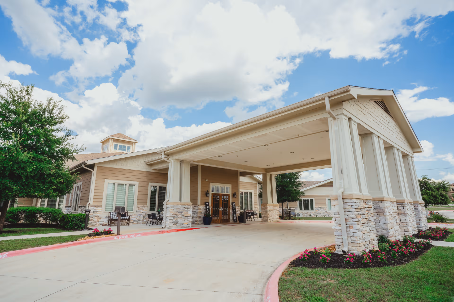 Exterior view of a senior living facility entrance with a covered drop-off area supported by stone and white pillars, surrounded by landscaped greenery and flowers under a partly cloudy sky.
