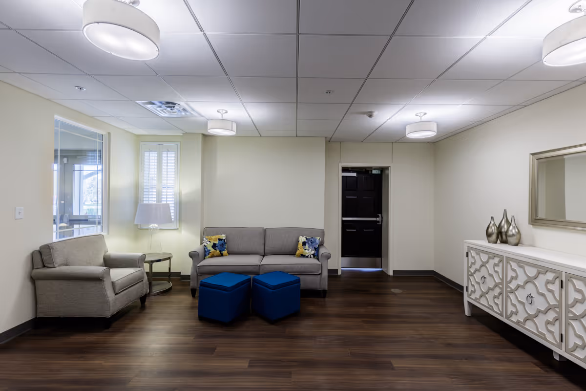 A modern living room area with a gray sofa adorned with floral pillows, a matching gray armchair, two blue ottomans, a round side table with a lamp, a white decorative cabinet with three metallic vases, and a large mirror above the cabinet. The room has wood flooring, light-colored walls, and ceiling lights.