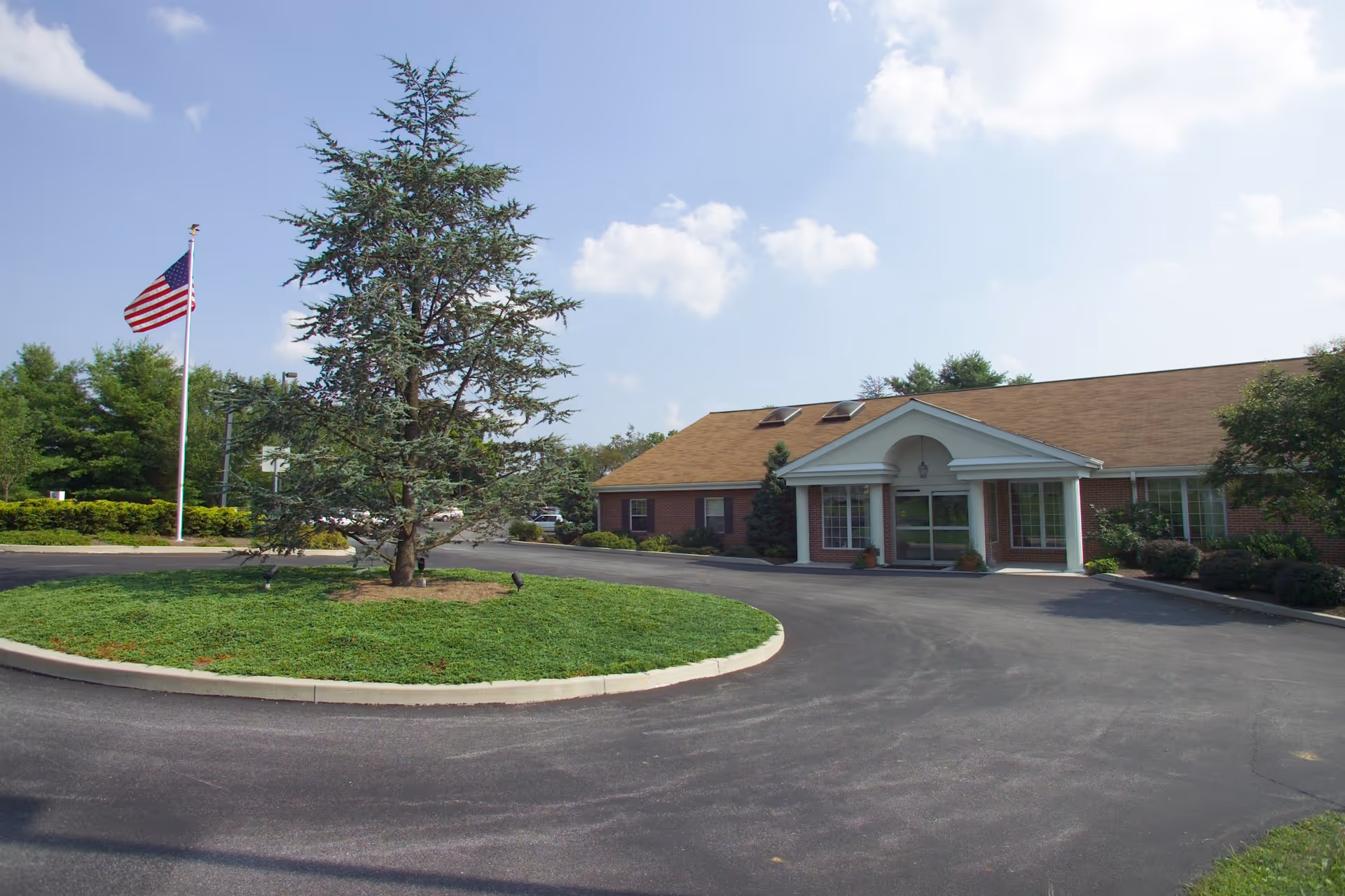 Single-story brick building with a columned entrance, circular driveway, landscaped island and an American flag on a flagpole.