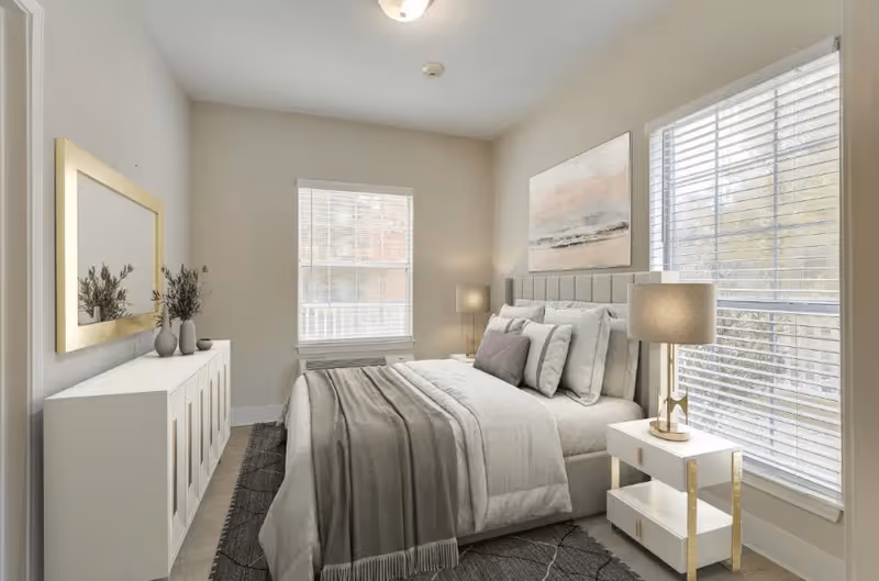 A bright and cozy bedroom with a neatly made bed featuring multiple pillows and a gray throw blanket. The room has two large windows with white blinds, a white nightstand with a lamp, a white dresser with decorative vases, and a large mirror above the dresser. The walls are painted light beige, and there is a piece of artwork hanging above the bed.