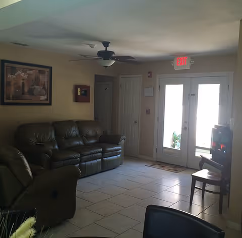 Interior view of a living room area with a brown leather sofa and recliner, a ceiling fan with light, a framed picture on the wall, double glass doors with an exit sign above, and a small TV on a stand with a chair beside it.
