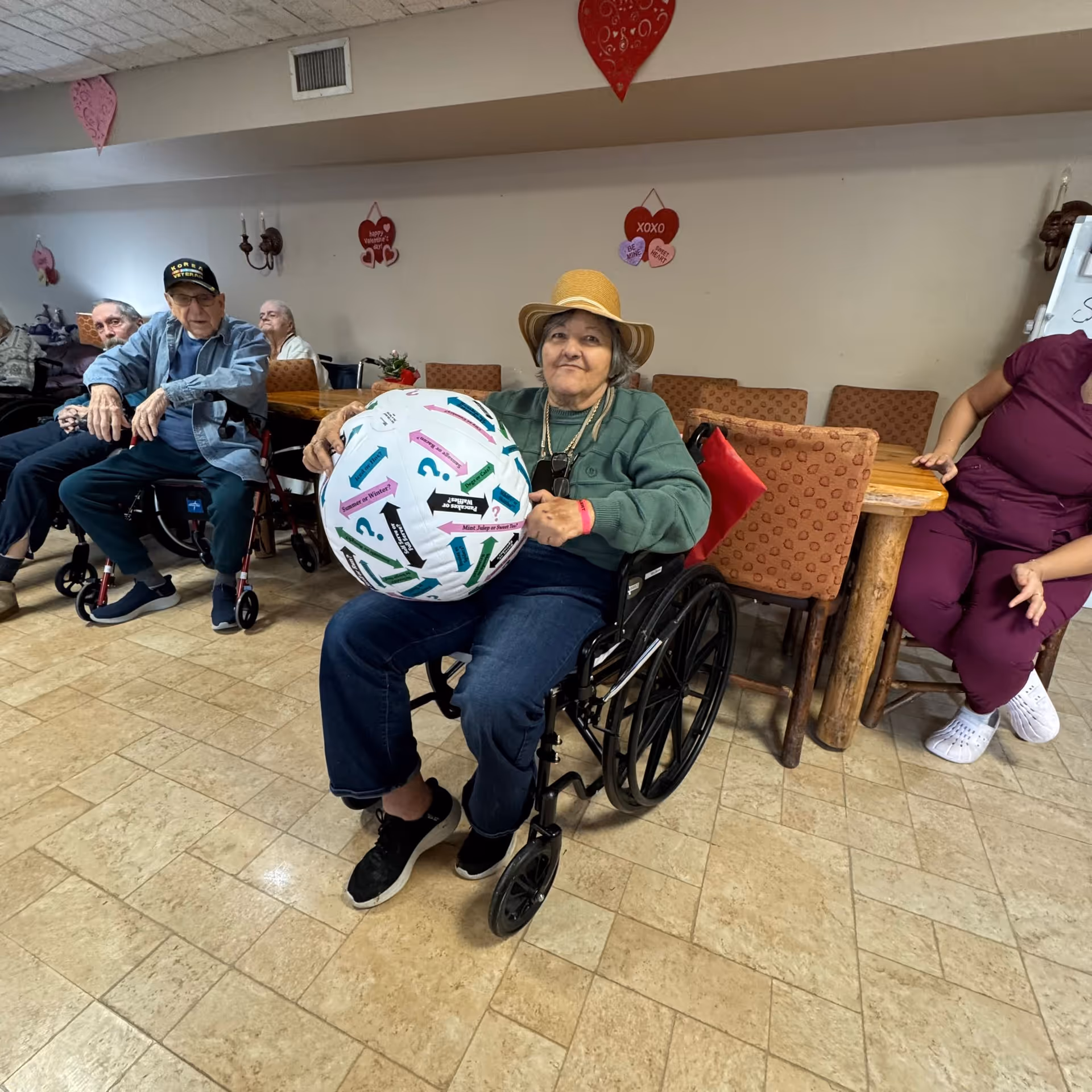 A group of elderly people sitting in a common room at a senior living facility. One woman in a wheelchair wearing a hat is holding a large inflatable ball with colorful arrows and text. Other residents are seated around a wooden table with cushioned chairs. The room is decorated with heart-shaped Valentine’s Day decorations on the walls.