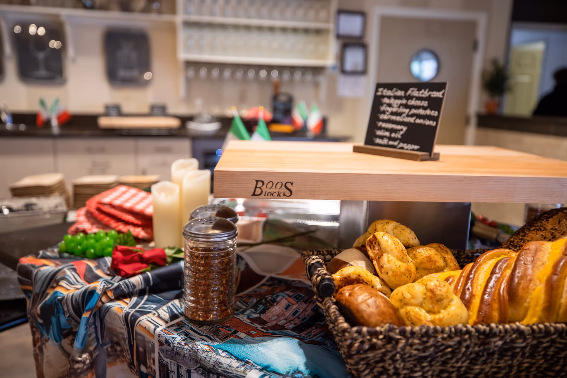 Buffet table in a communal dining area with baskets of bread and pastries, a wooden cutting board, condiments, and decorative candles.