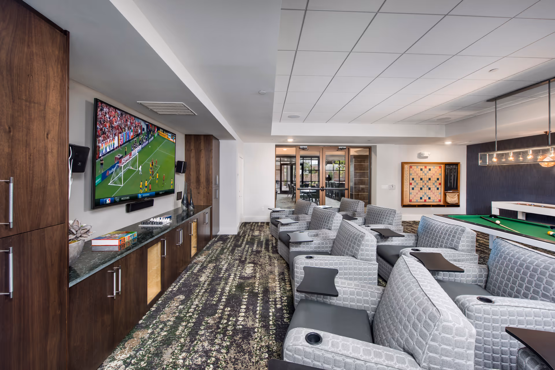 A modern entertainment room with multiple gray armchairs equipped with small black tables and cup holders, facing a large flat-screen TV mounted on a wall. The TV is showing a soccer game. The room has a patterned carpet, wooden cabinets beneath the TV, and a pool table and large Scrabble board on the far wall. Glass double doors lead to another room with tables and chairs.