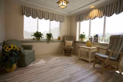 Sunlit senior living lounge with chairs, a small table, and potted plants by large windows.