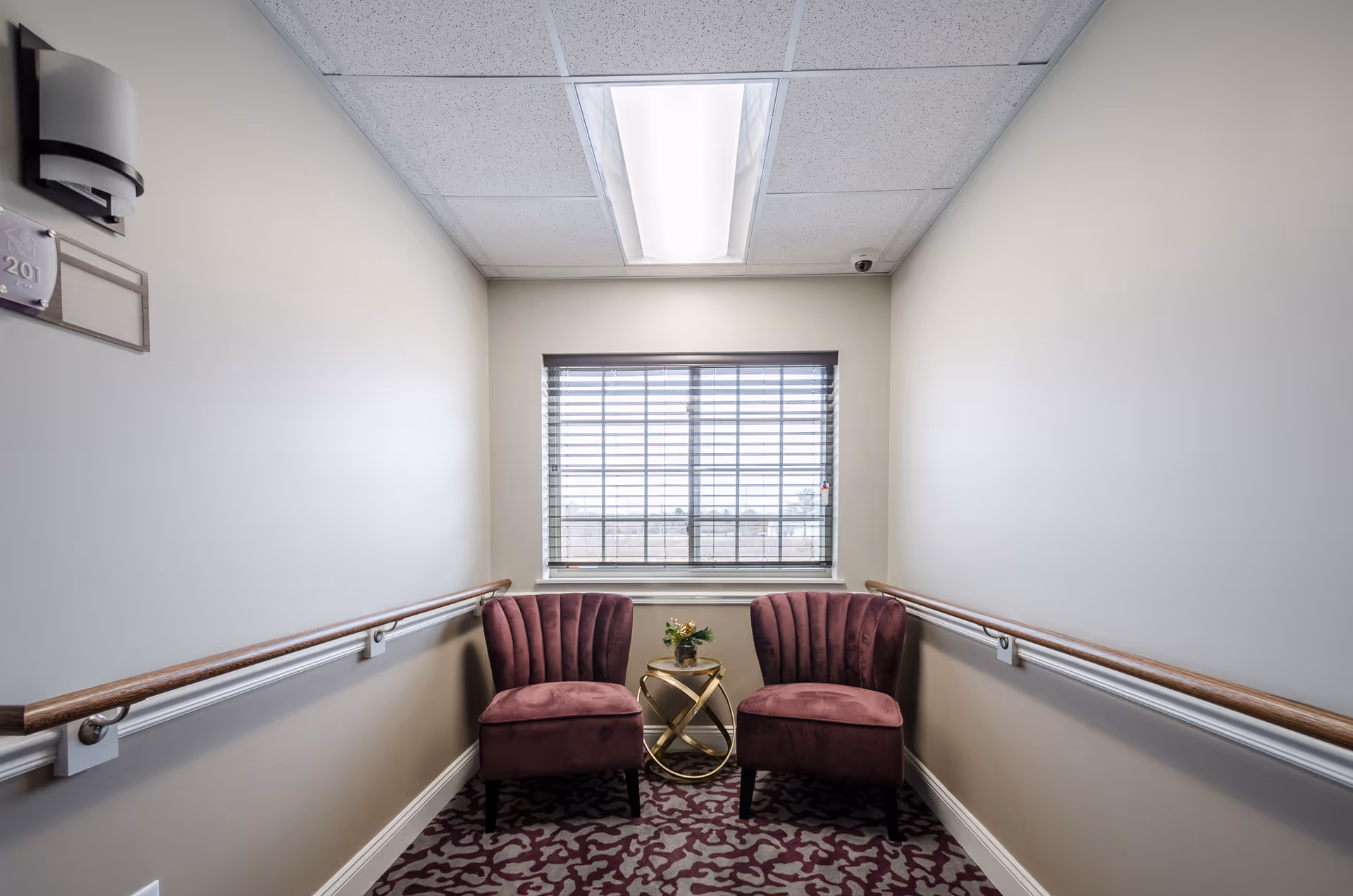 A small seating area in a hallway with two purple upholstered chairs and a small round gold table with a flower arrangement between them. The walls are beige with wooden handrails on both sides, a window with blinds at the end, and a ceiling light above.