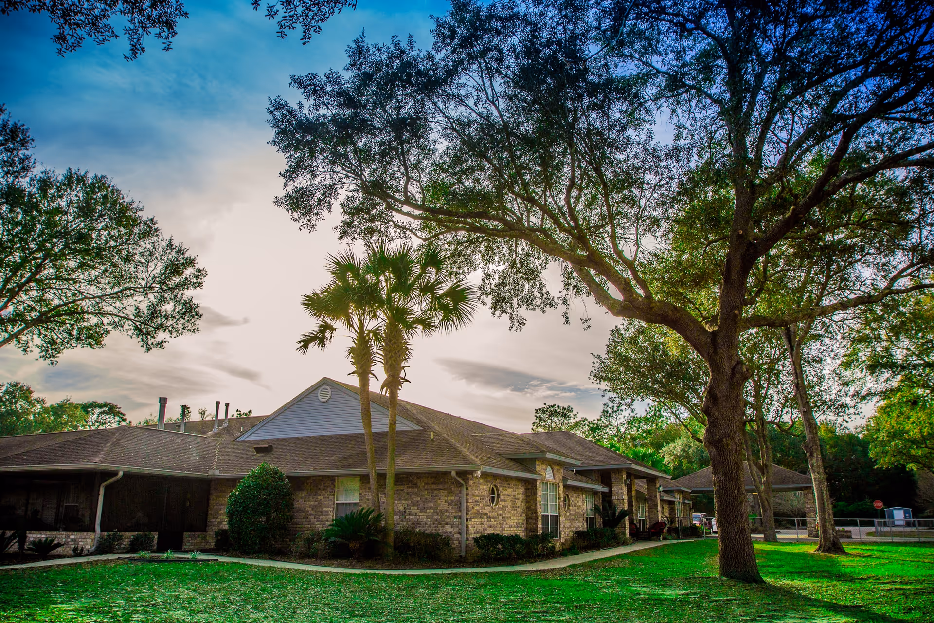 Exterior view of a single-story brick building with a sloped roof surrounded by green grass and large trees under a partly cloudy sky.